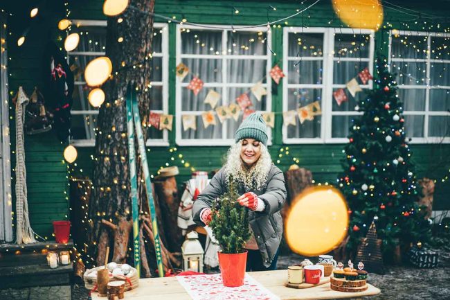A woman decorating a small tree in a pot with christmas decorations in the background.