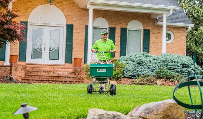 st-louis-lawn-care Man pushing a seedspreader machine over a grassy lawn infront of a large house.